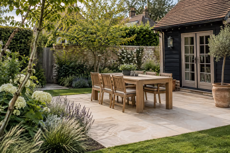 Rectangular pale limestone dining terrace with sturdy oak table and six coherent chairs beside modest garden house, planted border