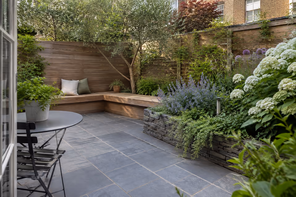 Small slate-gray stone backyard patio with built-in low timber bench along a fence, raised flowering border, coherent tight urban space